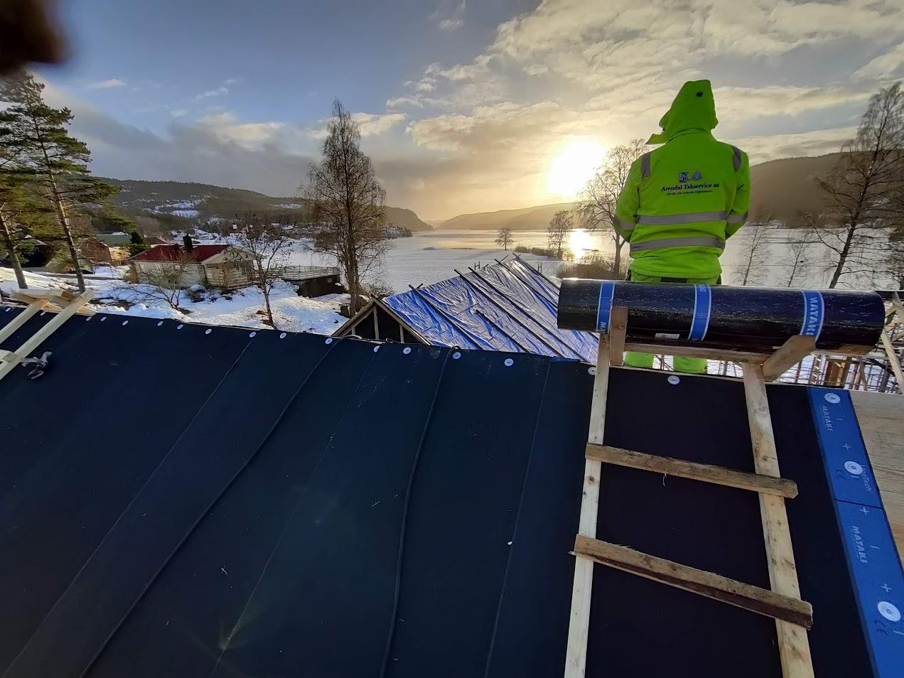 Taktekker i refleksjakke på et ferdig tak i morgenlys, med sjøen bak